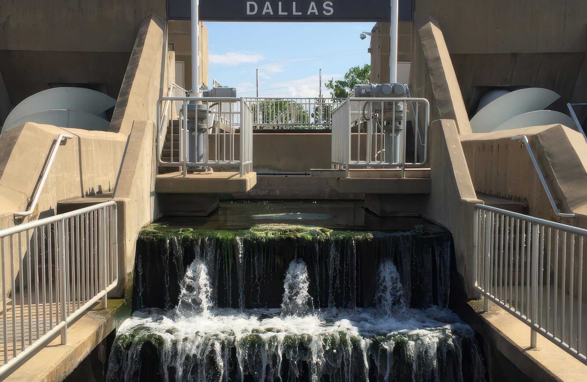 Water flows down algae-covered steps at a Dallas wastewater plant, flanked by Archimedes screw pumps and concrete structures.