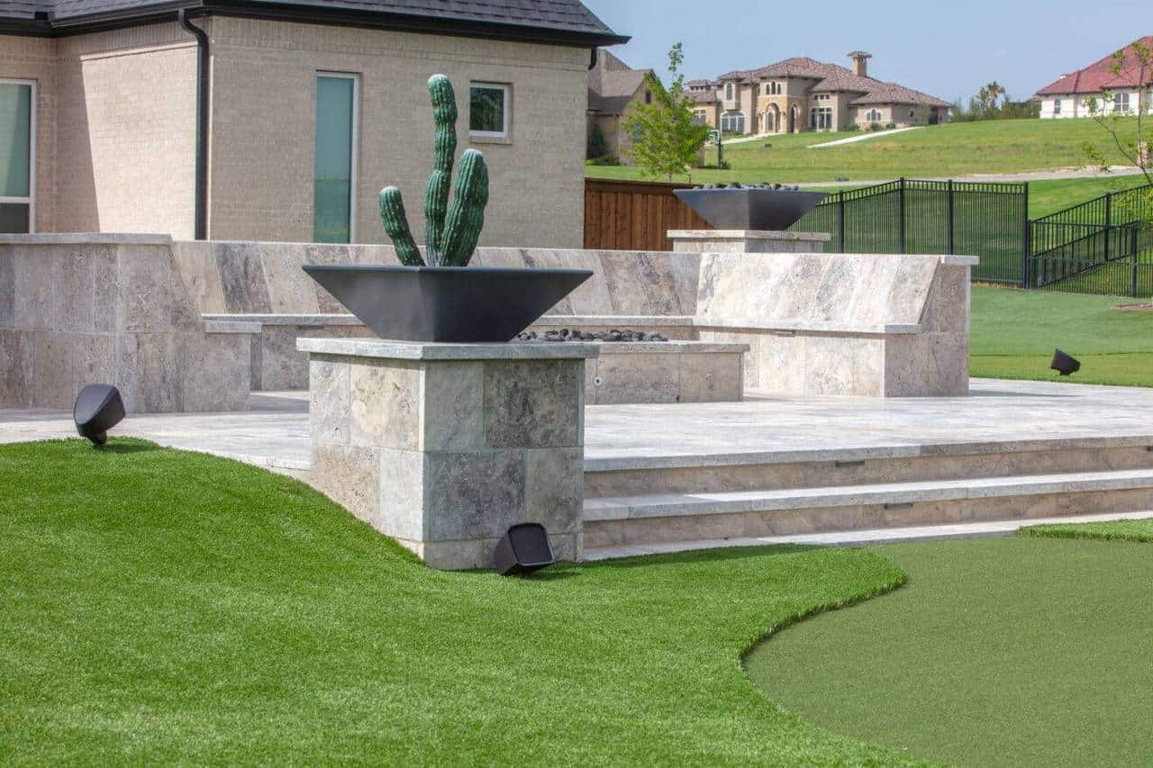 Modern backyard with stone patio, cactus in planter, and artificial grass, framed by luxury homes and manicured lawns.