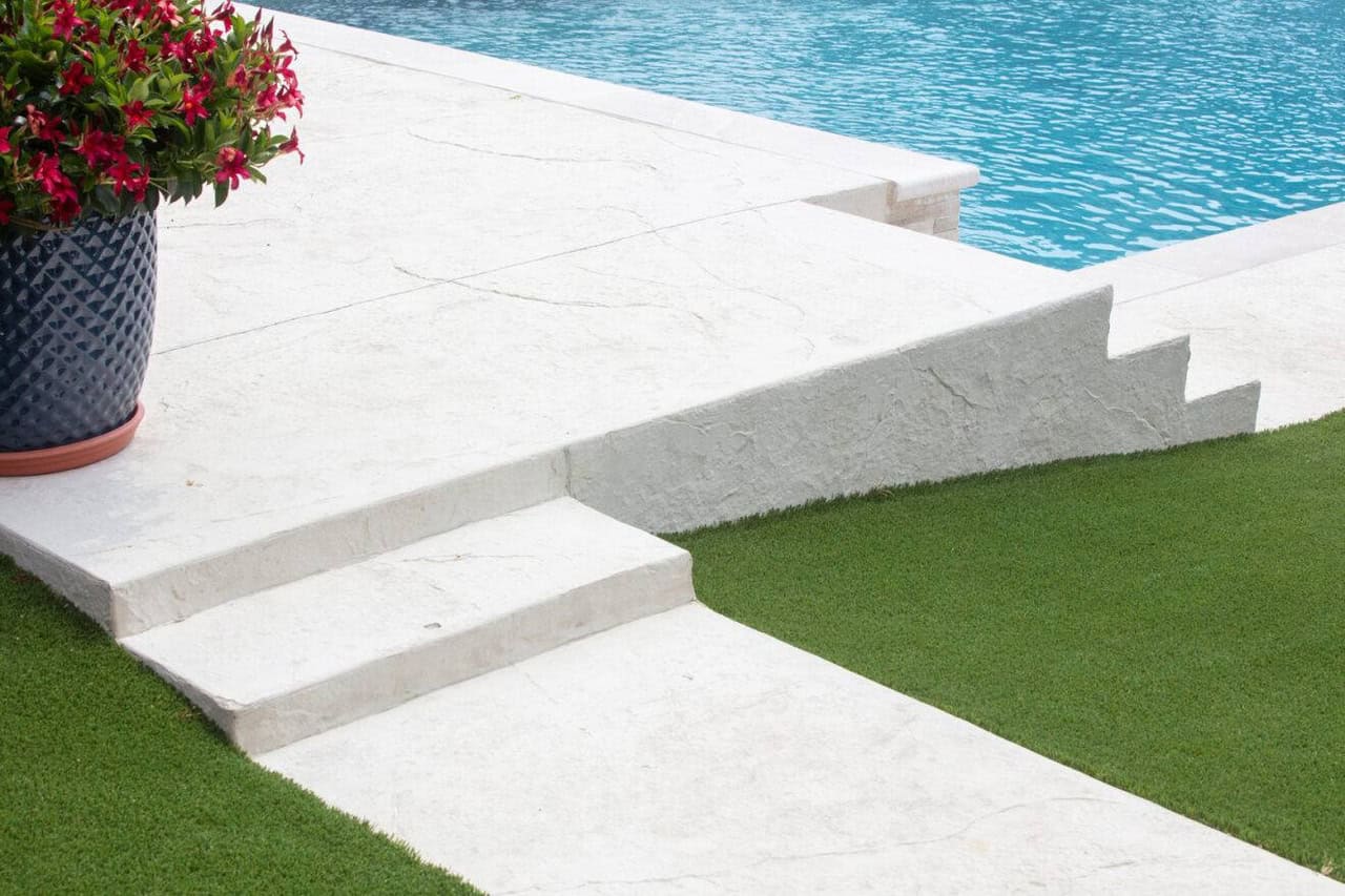 Artificial grass borders white stone steps leading to a pool, with a potted plant on the patio.