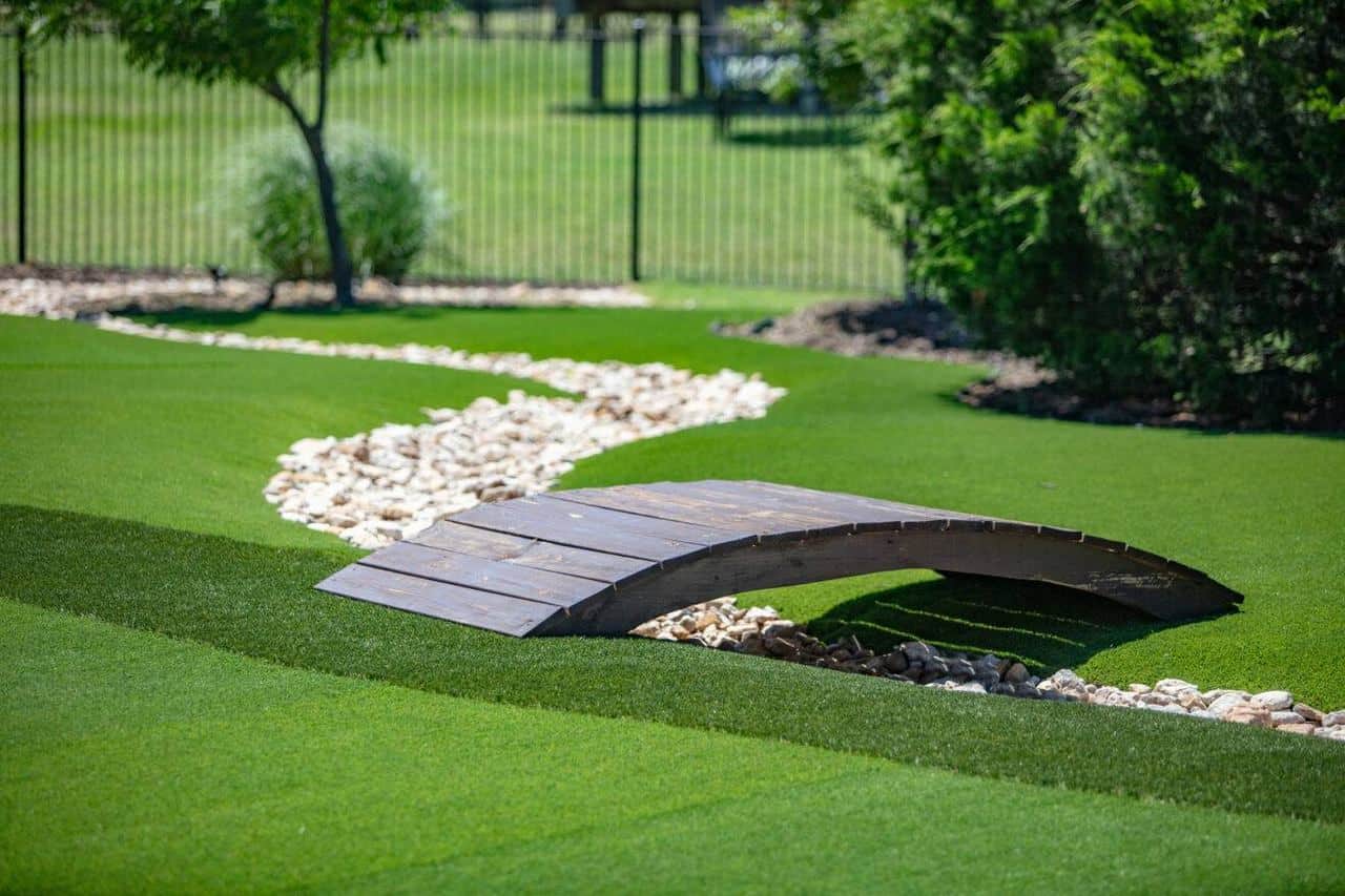 Wooden footbridge over a dry rock stream on a manicured artificial lawn, bordered by trees and a black metal fence.