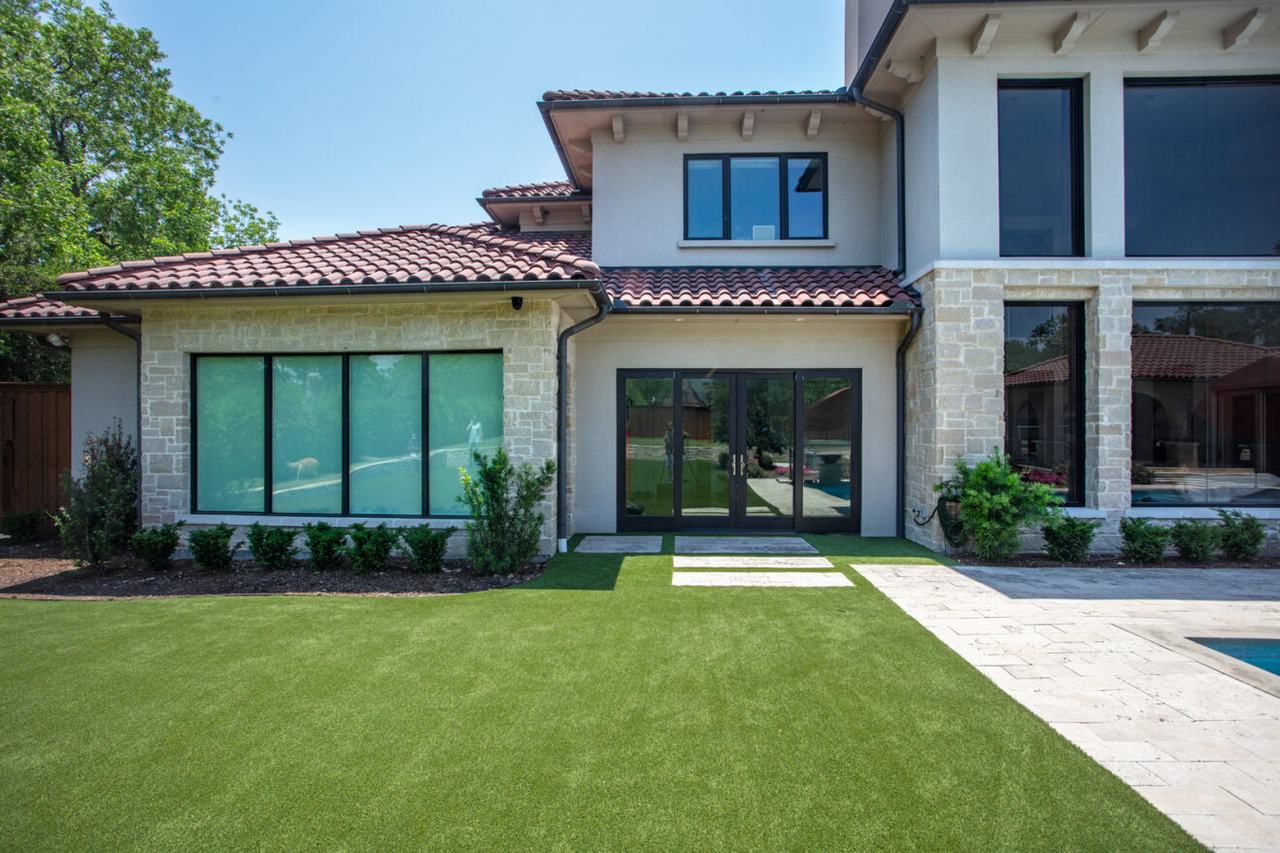 A modern home with large reflective windows faces a yard of artificial grass, where intense sunlight can cause turf to overheat and melt.