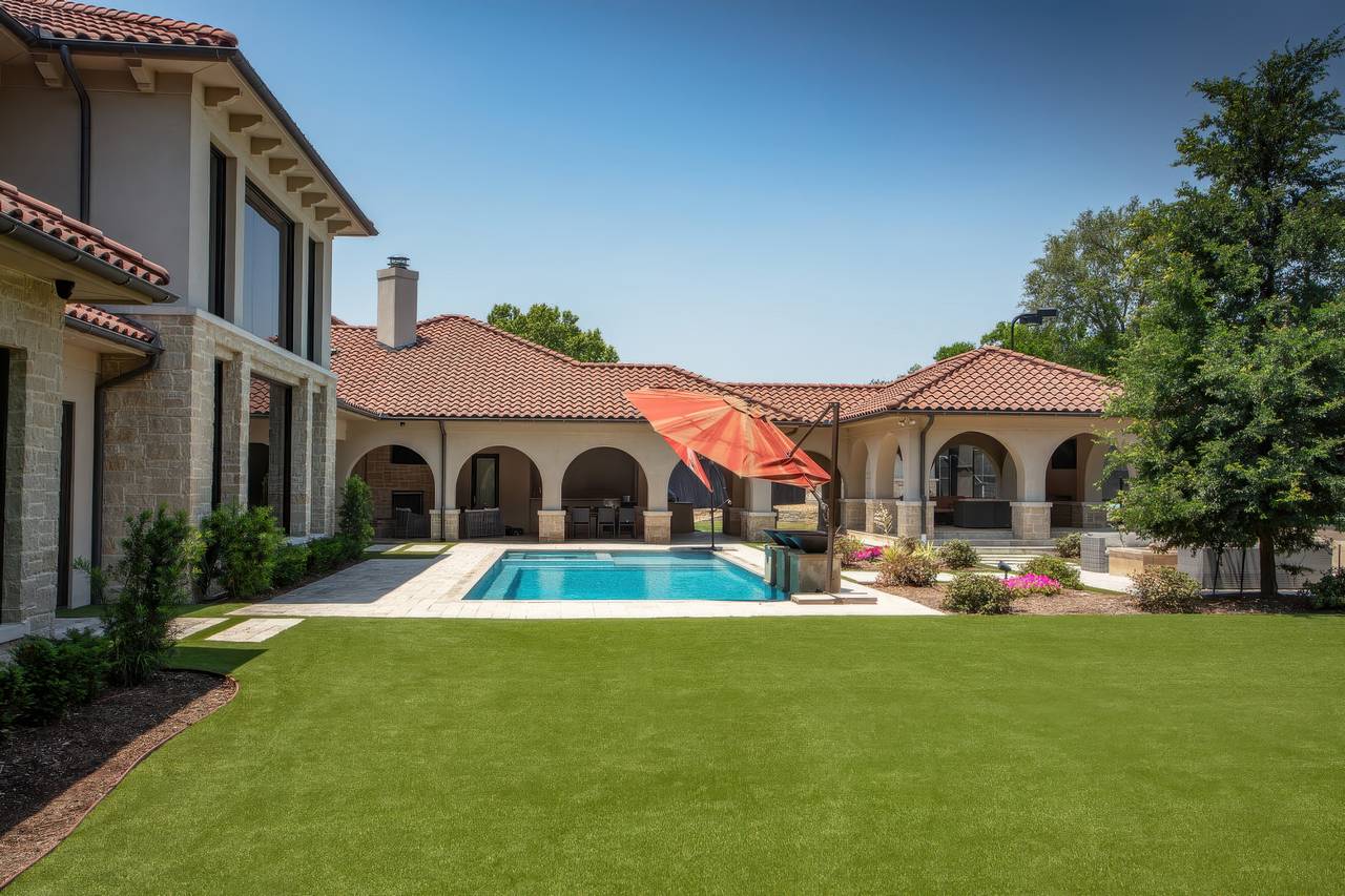 A luxury home with red tile roofing surrounds a backyard pool, featuring a lawn of artificial grass and a melted red umbrella.