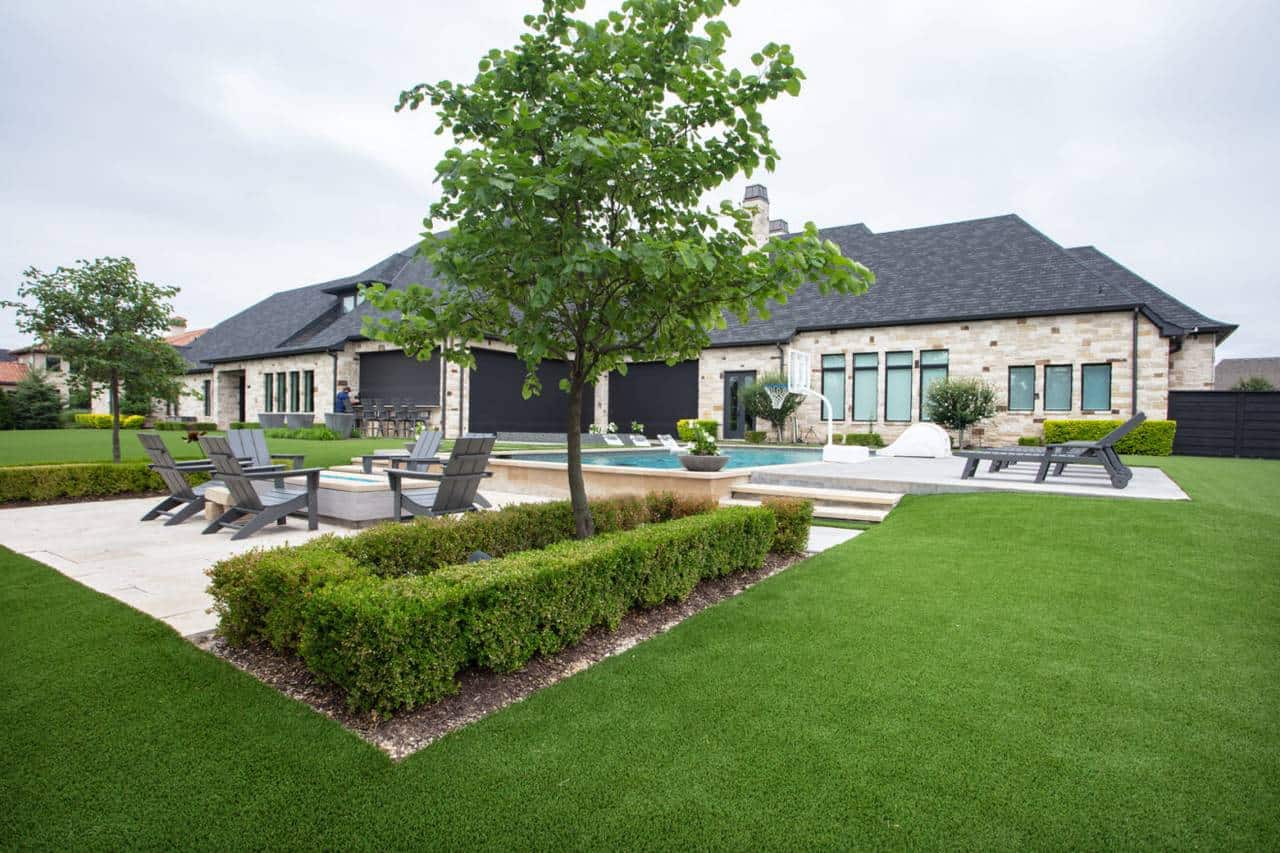 Modern backyard with artificial turf, patio seating, pool, and trimmed hedges in front of a large stone house.