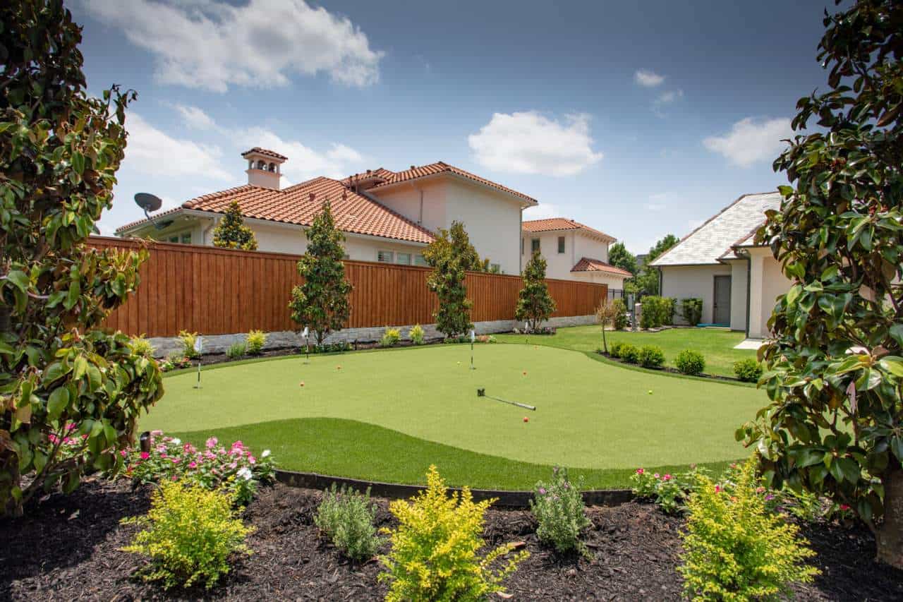 Backyard putting green with artificial golf turf, surrounded by plants, trees, and houses with red tile roofs.