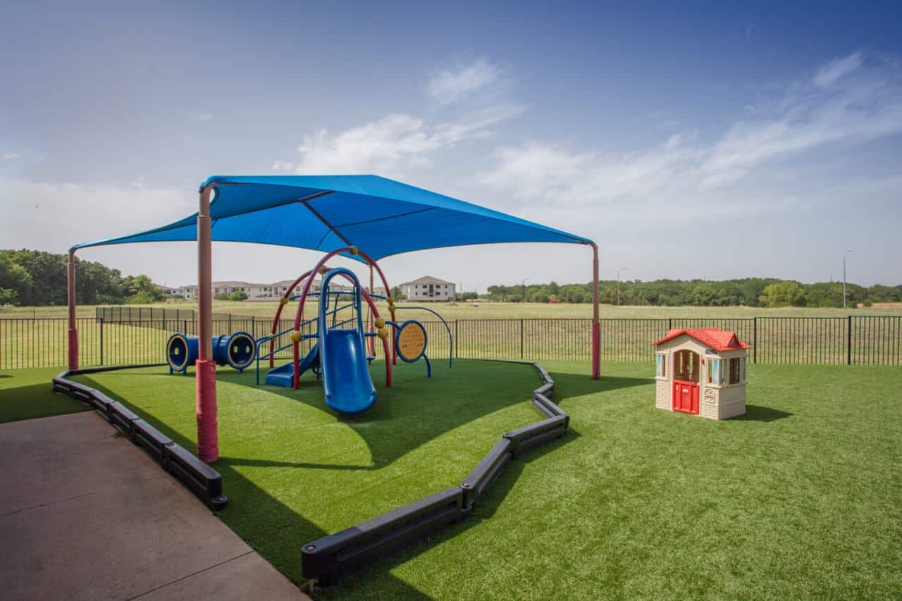 Playground with slide and shade canopy on artificial turf, next to a toy playhouse, enclosed by a fence in open grassy area.