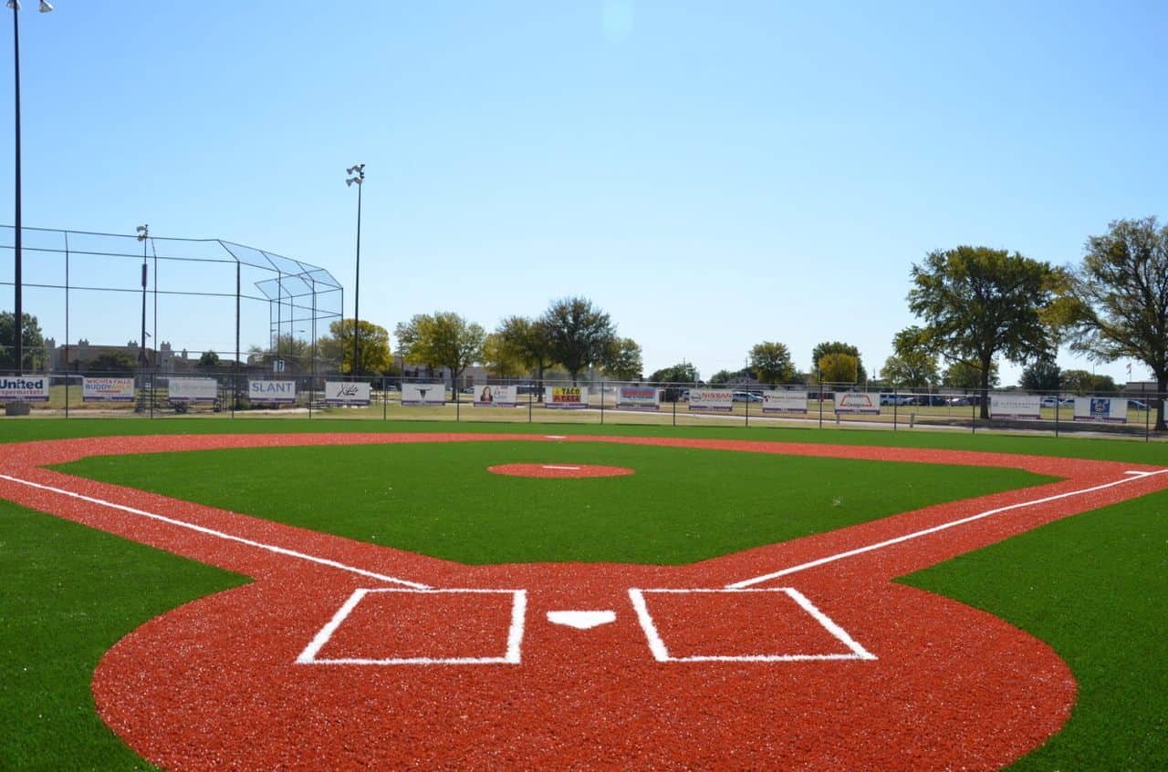 Baseball field with red and green artificial turf, white baselines, and backstop fencing under a clear blue sky.