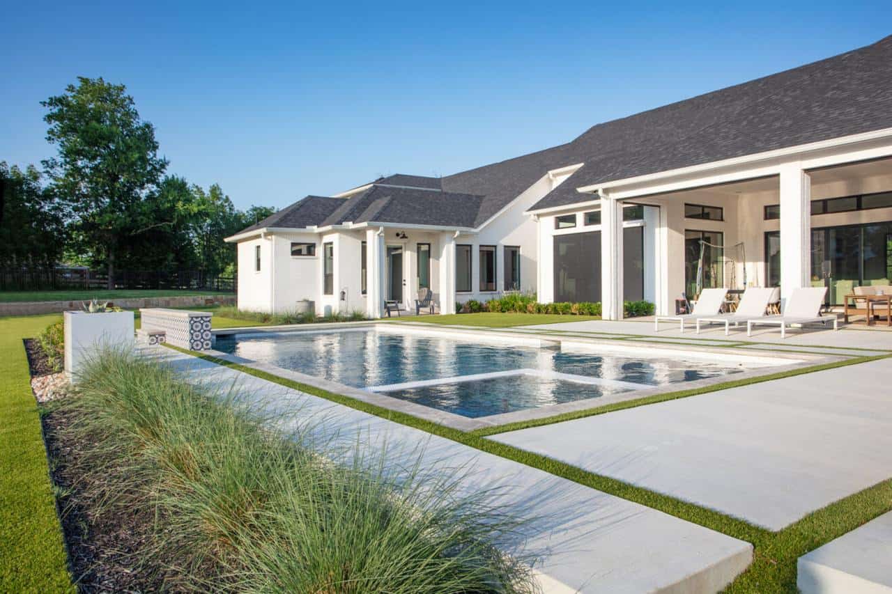 Modern backyard with pool, white lounge chairs, and artificial turf strips between concrete pavers beside a white house.