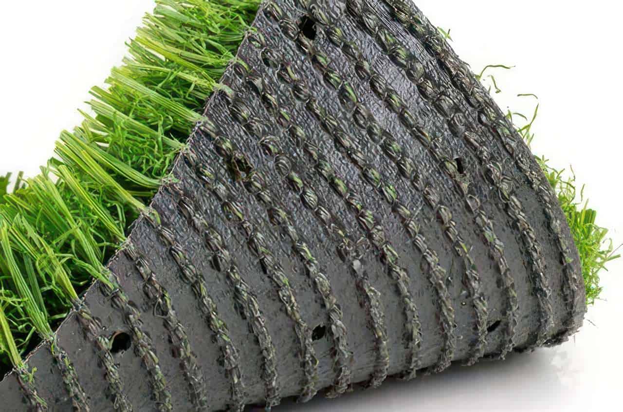 Close-up of artificial turf backing rolled to show drainage holes, stitched rows, and green synthetic grass blades on white surface.