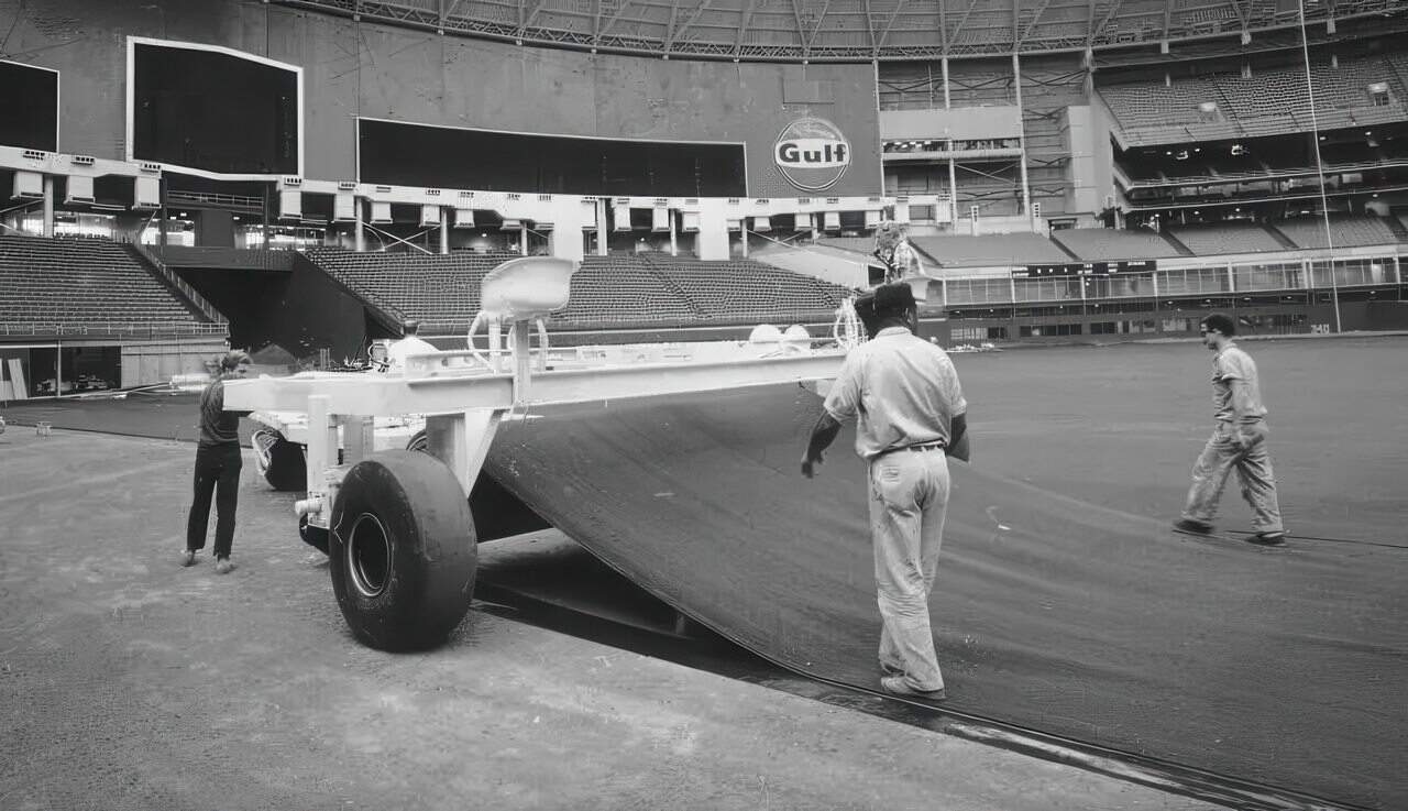 Workers unrolling early artificial turf in a large domed stadium, with vintage machinery and empty seating in the background.