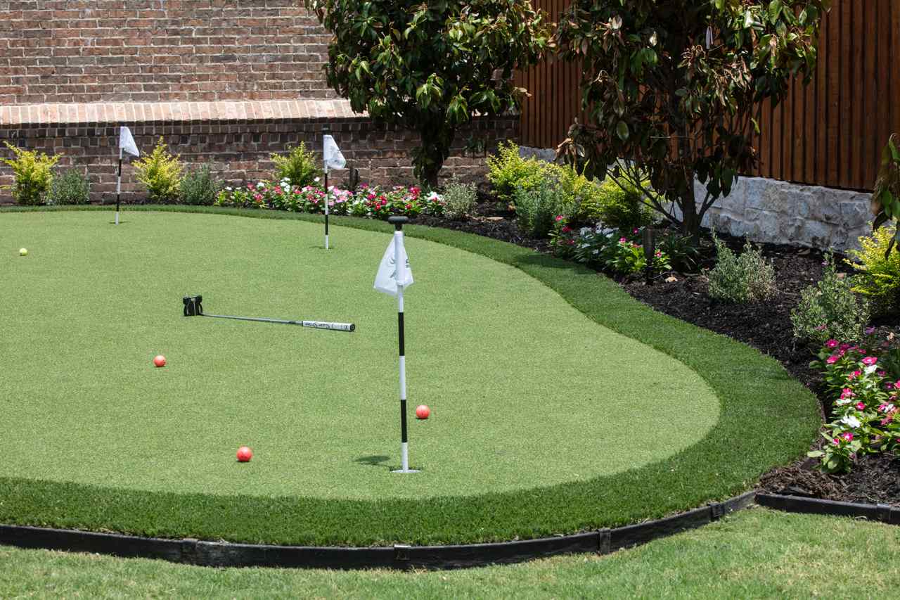 Backyard putting green with artificial grass, golf holes with flags, and surrounding flowers and trees.