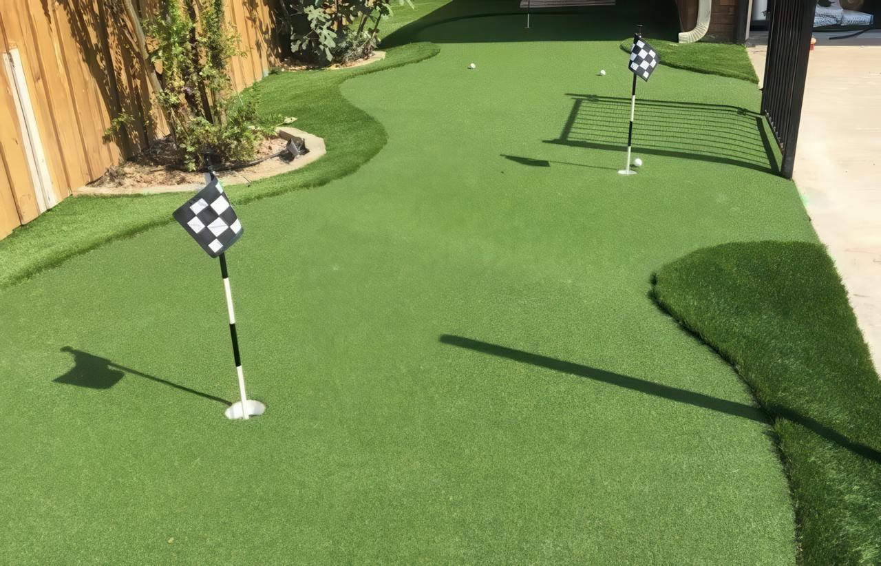 Artificial putting green in backyard with checkered flags, golf balls, curved edges, and surrounding natural landscaping.